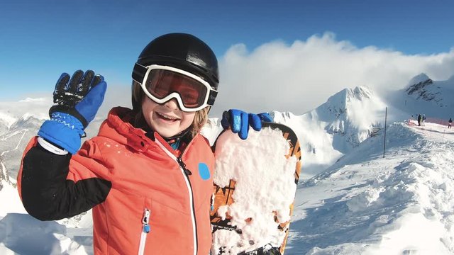 Smiling child with snowboard waving hand on snow mountain background. Enjoying winter vacation on ski slopes in sunny day. Having fun with outdoors activity, sport