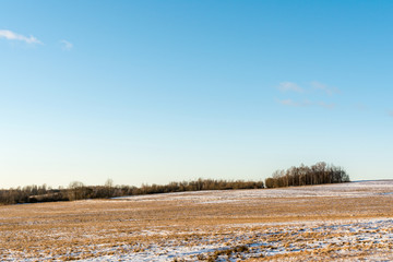 View of a hilly horizon with a snow-covered field with orange dry grass and shrubs. Clear blue orange sky in early spring in evening