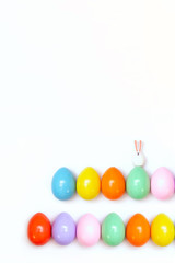 Obraz premium Top view of an Easter composition of painted eggs in bright juicy colors on a white background with a little rabbit. Holiday concept, flat layout, minimalism
