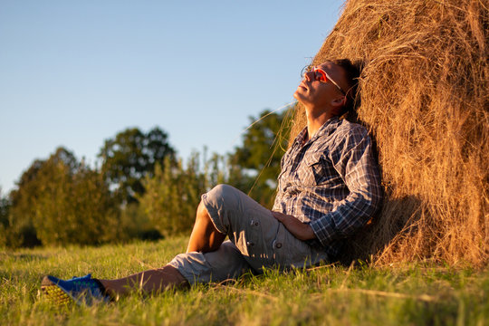 A man in a field sitting on a haystack at sunset.