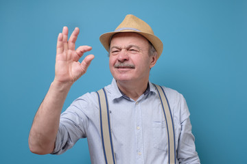 Naklejka premium Caucasian senior man with mustache wearing summer hat show small size by fingers demonstrate tiny measure, tell about little decreased prices. Studio shot on blue wall