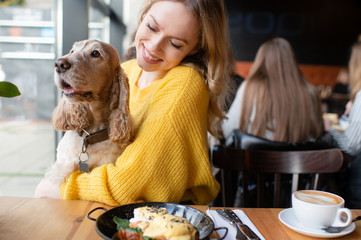 Young caucasian cheerful blonde girl sitting and hugging together with her lovely cocker spaniel dog in caffe at the table with lunch meal and cappuccino.