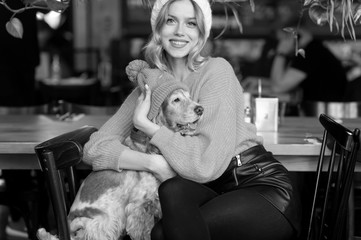 Positive smiling and happy blonde girl in yellow sweater and white hat hugs her kind cocker spaniel...