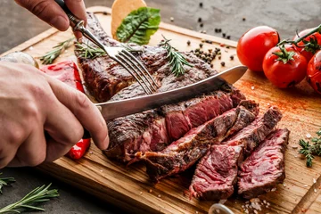 Gartenposter Hands cut grilled tomahawk meat medium rare or rib eye steak on wooden cutting board with grilled vegetables on dark background, close up © Jukov studio