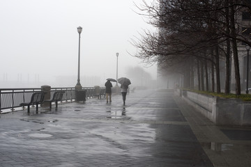Moody image of two people walking in the fog and rain holding umbrellas in Jersey City across from the New York City skyline.