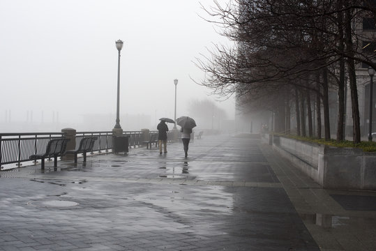 Moody image of two people walking in the fog and rain holding umbrellas in Jersey City across from the New York City skyline.