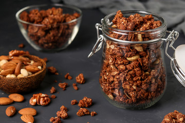 Granola crispy muesli with natural honey, chocolate and nuts in a glass jar and bowl against a dark background, healthy food, Closeup