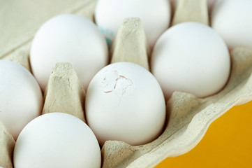 package with a dozen white eggs on a yellow background, one egg broken