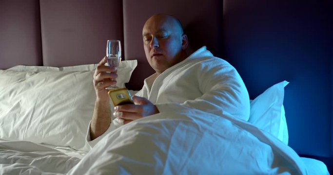 Adult Man Is Relaxing Before Sleep, Lying In Bed With Glass Of Wine And Smartphone