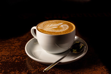 A Cup of cappuccino with a pattern on the foam is on the table in the early morning, while it is still dark.