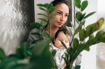 Pretty woman with black hair with her pretty little child sits on a windowsill behind the green flower