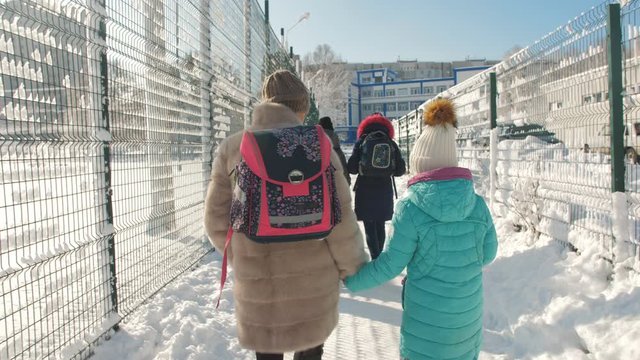 Mother and young daughter in the winter go to school together. Mother brings daughter's backpack on his shoulder.