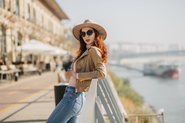Shapely caucasian young woman in hat standing near sea and looking to camera. Outdoor photo of positive ginger girl waiting friend at embankment.