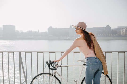 Portrait from back of lovable ginger girl touching her bicycle. Outdoor photo of fascinating slim woman standing on bridge and looking at river.