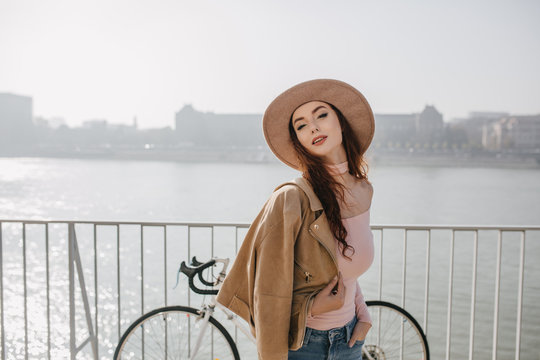 Carefree ginger woman in beige jacket posing near bicycle. Outdoor photo of pleased red-haired girl enjoying good day at sea embankment.