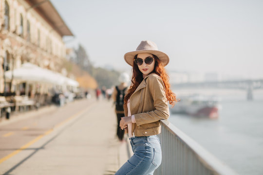 Slim red-haired girl in trendy hat standing in confident pose near sea. Outdoor portrait of magnificent white female model in casual attire enoying good weather.