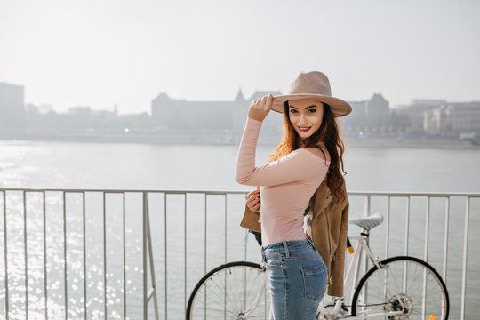 Outdoor photo of graceful slim woman in jeans touching her hat, standing on bridge on city background. Smiling active girl with bicycle enjoying river views.