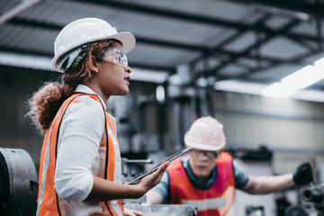Female industrial engineer wearing a white helmet while standing in a heavy industrial factory behind she talking with workers, Various metal parts of the project - Powered by Adobe