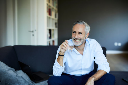 Smiling Mature Man Drinking Glass Of Water On The Sofa At Home