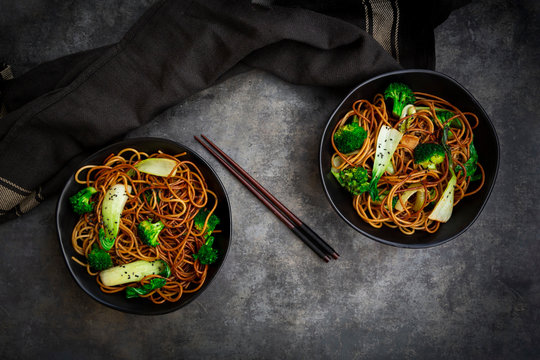 Overhead view of two bowls of soba noodles with pak choi and broccoli, soy sauce and black sesame