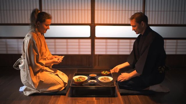 Young couple seiza sitting on floor pillows with Japanese ryokan kaiseki dishes, eating soba noodles, natto, edamame and soy sauce on table by sliding paper doors