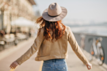 Blur portrait from back of graceful curly woman in leather jacket walking down the embankment. Outdoor photo of female model in casual outfit enjoying good day in european town.