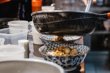 Selected focus view, pouring Thai style brown soup with meat, pork and meat ball onto rice noodle and vegetable in bowl, on counter at restaurant's kitchen.