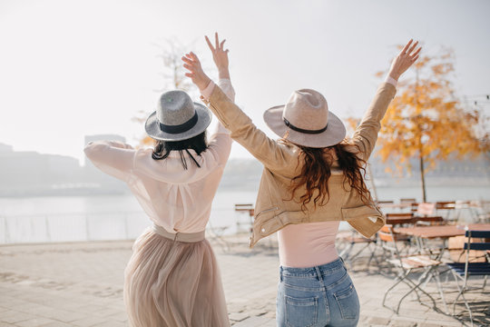 Portrait from back of two excited ladies expressing positive emotions enjoying river views. Photo of girls in hats jumping in outdoor cafe in sunny autumn day.