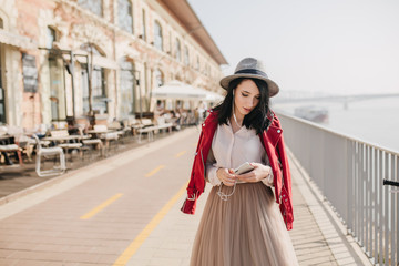 Well-dressed young woman in hat walking down the street in her leisure time. Fashionable brunette girl with phone in hat posing at embankment in sunny morning.