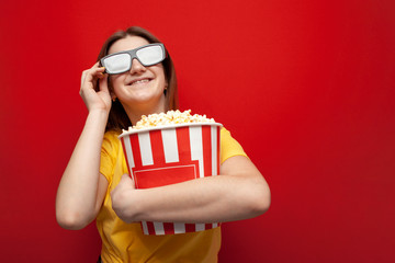 happy hipster girl with popcorn puts on 3D glasses and smiles on a red colored background