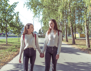 Two girls girlfriends schoolgirls 12-14 years old, walk in summer park, background road trees, hold hands, laugh, have fun talking, return after school, road home. Emotions of positive and relaxing.