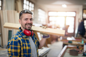Portrait of professional carpenter holding wood plank material on his shoulder and smiling....