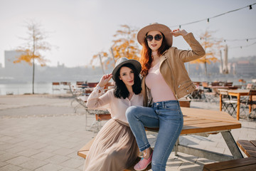 Blissful red-haired girl in jeans relaxing in street cafe with friend. Excited ladies posing together on autumn nature background.