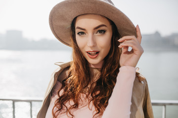 Enchanting brown-haired girl playfully posing on river background. Outdoor photo of joyful white female model in hat smiling on embankment in sunny day.