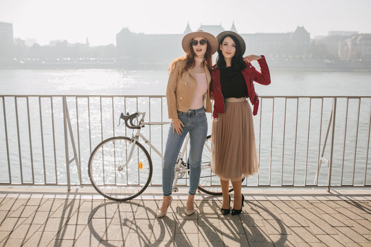 Full-length portrait of shapely white girl in jeans spending time with brunette sister. Outdoor photo of attractive woman in beige skirt having fun with ginger female friend.