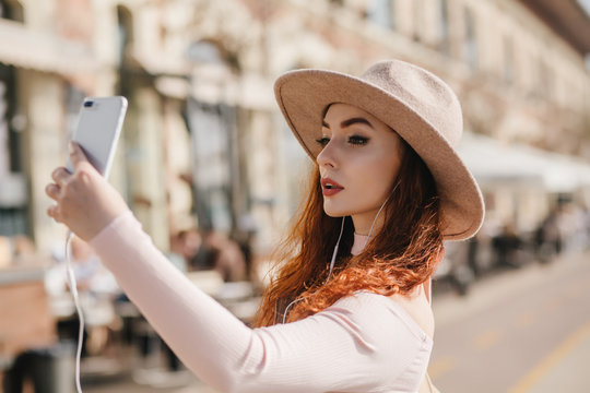 Serious white woman with dark makeup taking picture of herself. Outdoor photo of concentrated ginger lady using phone for selfie on urban background.