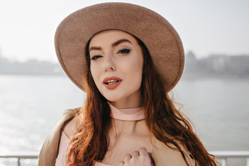 Close-up portrait of beautiful ginger girl isolated on sea background. Outdoor photo of cute red-haired young woman in hat spending time near river.