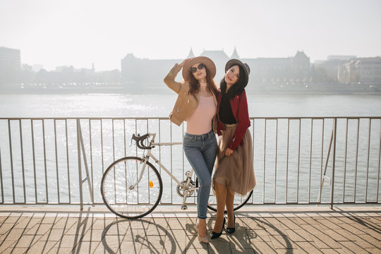 Full-length photo of graceful woman in jeans posing with brunette sister at embankment. Outdoor shot of two ladies standing beside bicycle on river background.