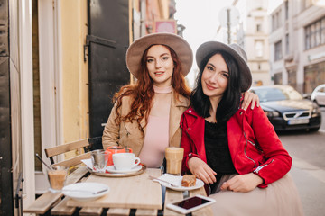 Refined red-haired girl embracing her female friend in cafe. Outdoor portrait of two sisters enjoying latte on street background.