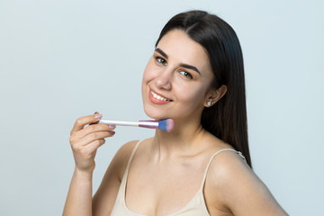 Close-up of a young girl in a light top on a white background making a facial make-up. A pretty woman holds a cosmetic brush near her face and smiles.