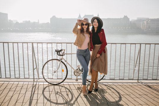 Full-length photo of charming brunette girl in skirt posing with female friend in jeans. Portrait of two stylish sisters standing near bicycle on the bridge.