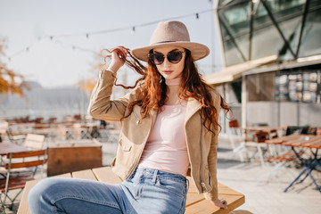 Carefree female model in dark glasses posing in hat in street cafe. Outdoor portrait of ginger serious woman in jacket enjoying morning.