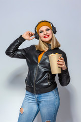 Portrait of Caucasian Female Blond Girl With Headphones On Holding Bucket of Popcorn. Posing in Leather Jacket and Yellow Hat Against White.