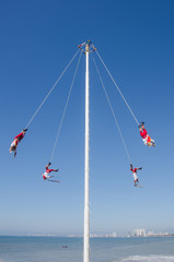 Four indigenous men swinging from ropes on a tall pole while playing native instruments.