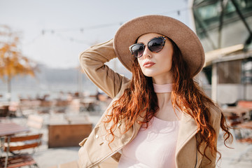 Pale woman with wavy dark hair looking up and touching her hat. Outdoor photo of chilling white girl in sunglasses posing on blur background in warm day.