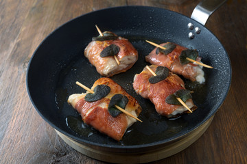 Roasted chicken saltimbocca wrapped with air-dried ham and sage leaves in a black pan on a rustic wooden table