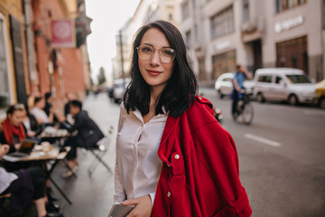 Cheerful caucasian girl with tired smile posing on city background. Outdoor portrait of black-haired glad young lady standing on the street.