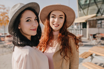 Fototapeta premium Brunette girl in hat playfully looking over shoulder while posing with friend. Ginger caucasian young woman spending leisure time with sister.