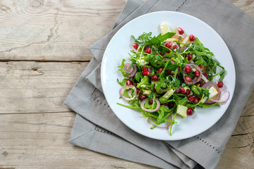 Arugula or rocket salad with pomegranate seeds, red onions and parmesan served on a white palte on a rustic wooden table, copy space, high angle view from above