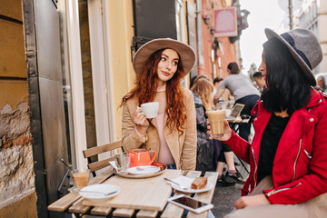 Elegant ginger girl drinking coffee an talking with friend. Outdoor photo of two ladies enjoying latte in weekend.